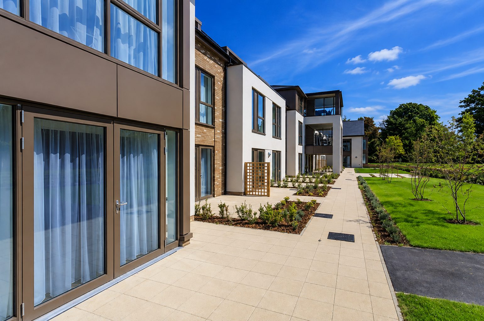 Beige-grey aluminium composite panels around glazed ground-floor garden doors at Liberham Lodge, with brick, white render and more panel-framed windows along the paved terrace