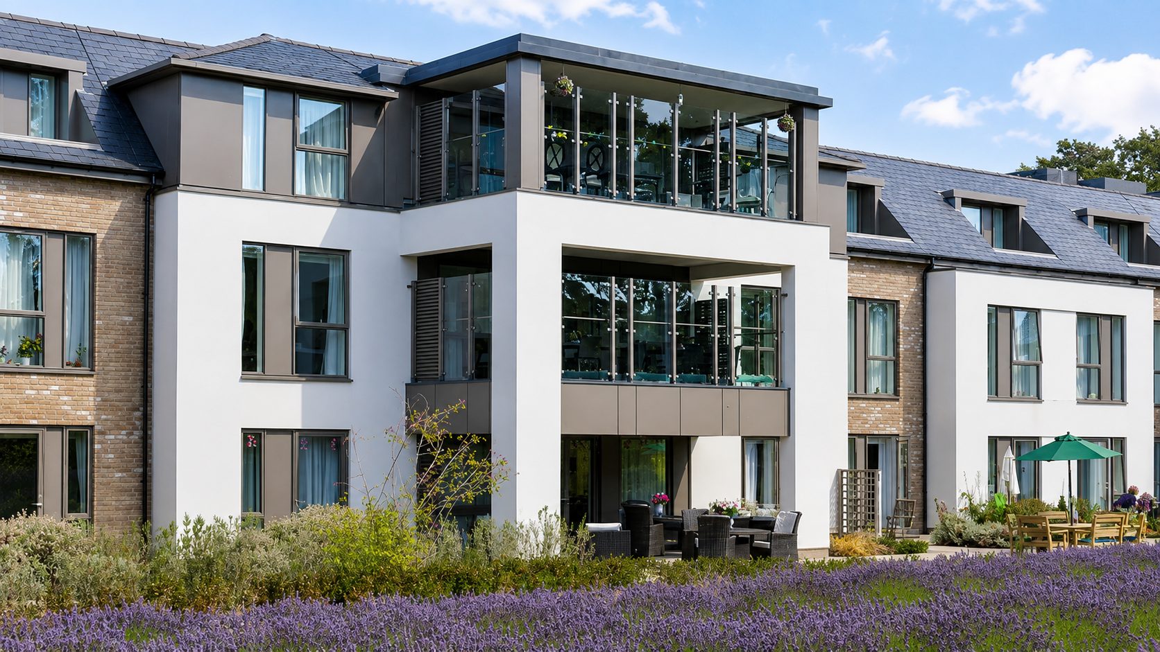 The central garden bay of Liberham Lodge, with glazed lounges on two floors, a beige-grey panel band beneath the upper glazing and panelled dormers in the slate roof either side