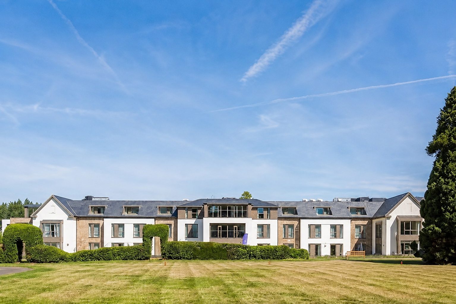 Liberham Lodge's long garden elevation across a striped lawn, with gabled end wings, a central glazed bay and panel-framed windows and dormers between white render and brick