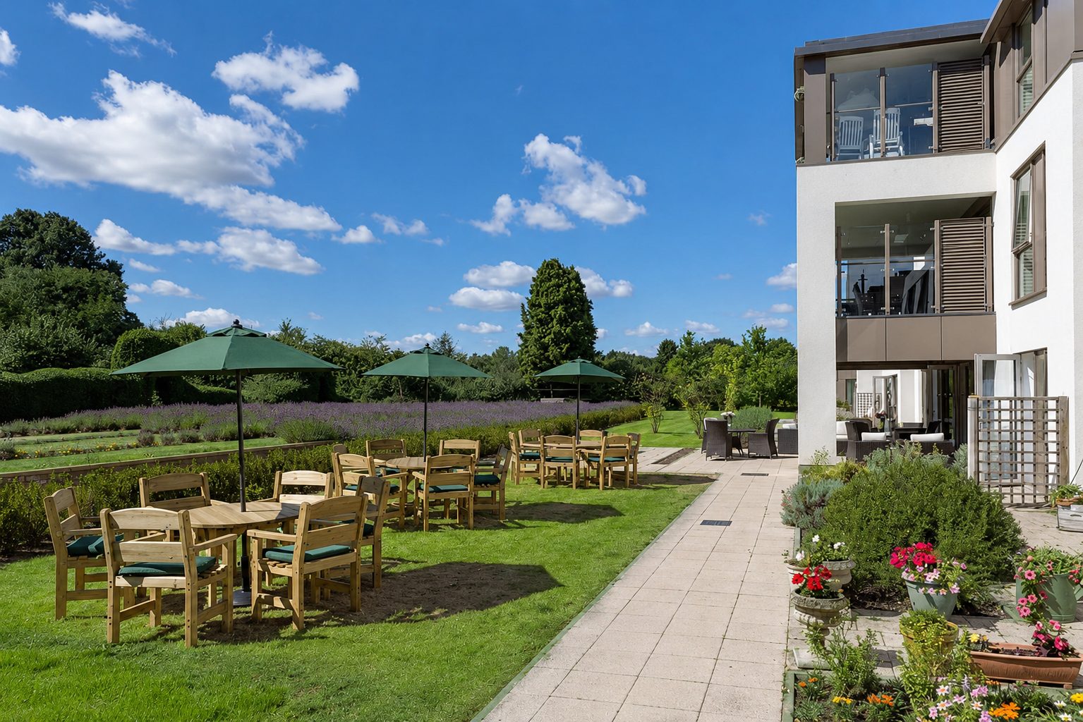 Wooden garden tables and green parasols on the lawn beside a paved terrace, below glazed lounges with a beige-grey panel band and louvred shutters