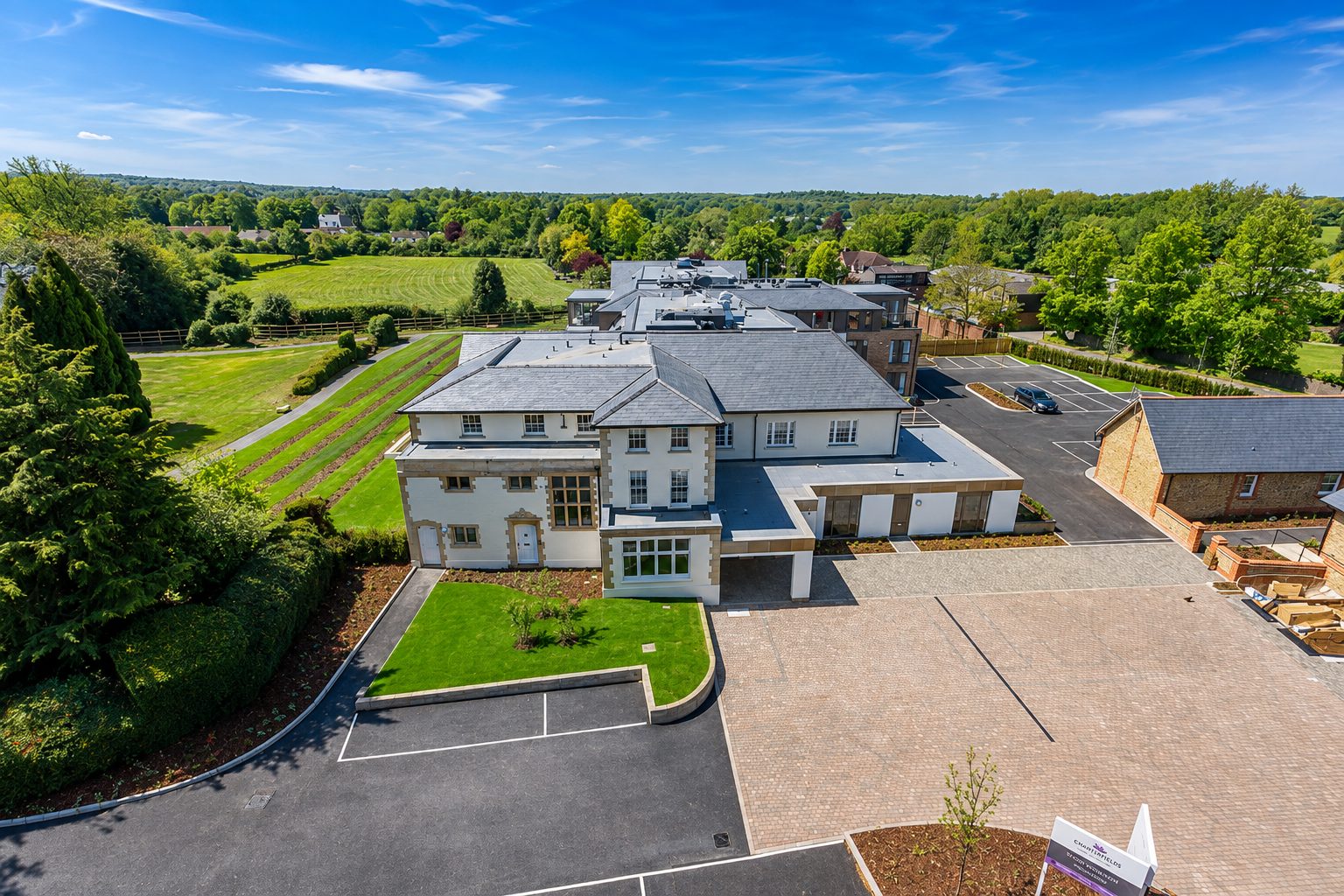 Aerial view of the retained rendered house with stone quoins, the entrance court and car park, and the new wing behind it among trees and fields