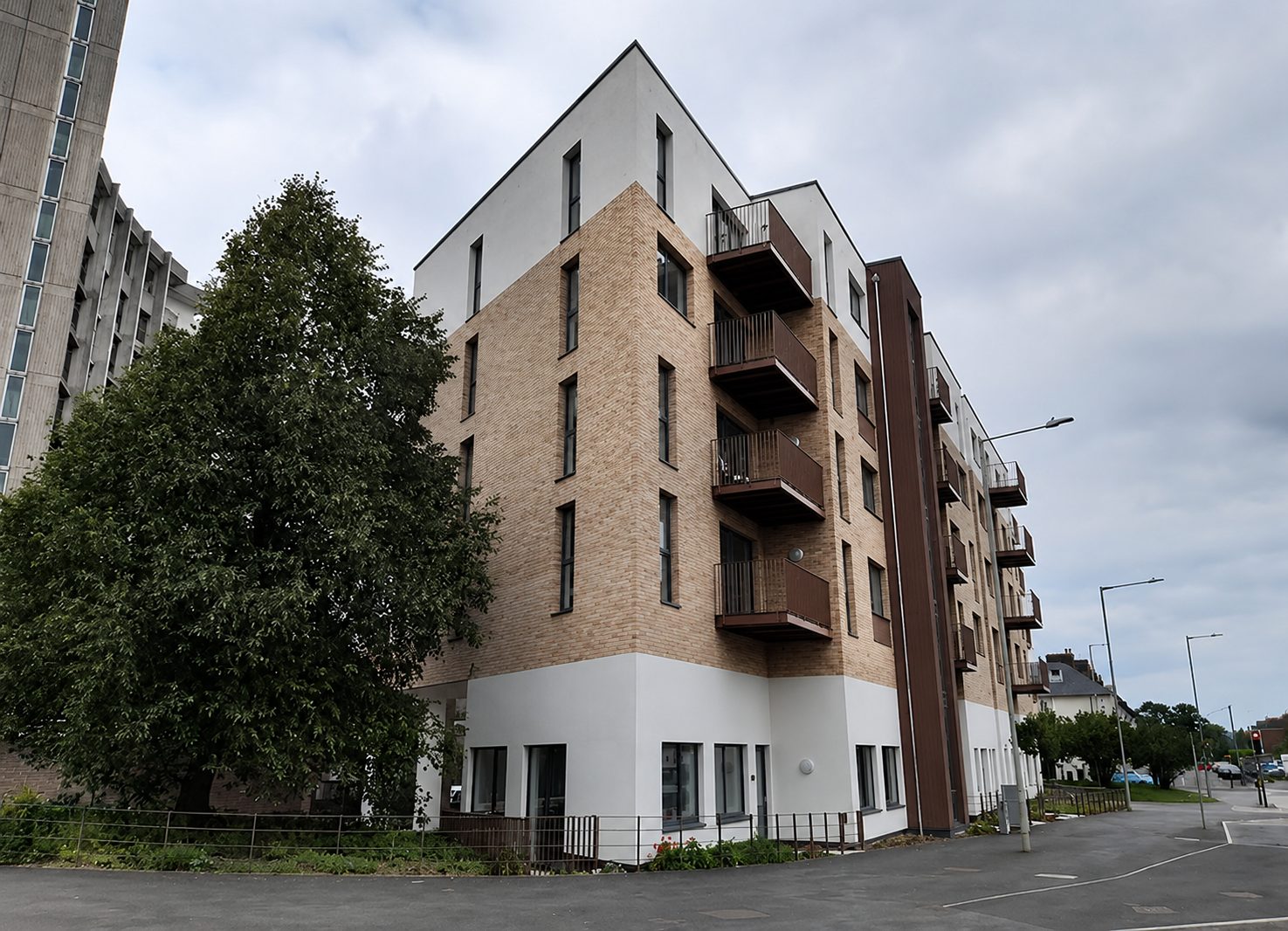 Corner of a six-storey apartment block in buff brick slip over a white-rendered ground floor, balconies stacked up one side, a tree in front