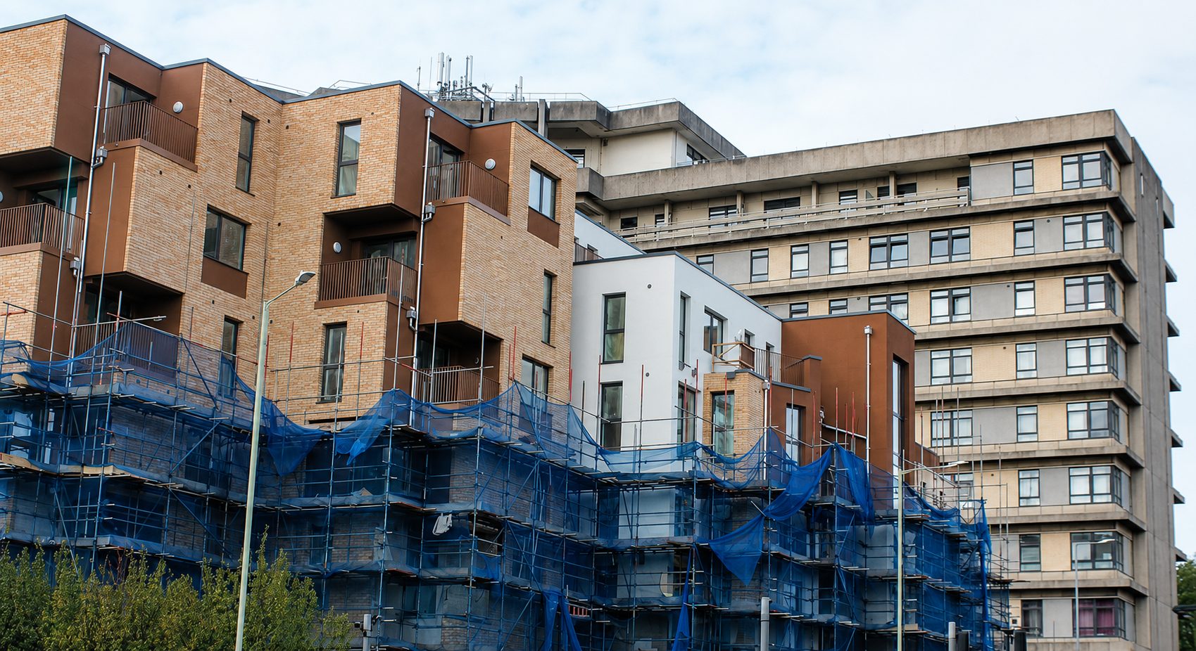 Upper storeys of a new wing finished in buff brick slip with brown bands, blue-netted scaffolding around the lower floors, a concrete tower behind