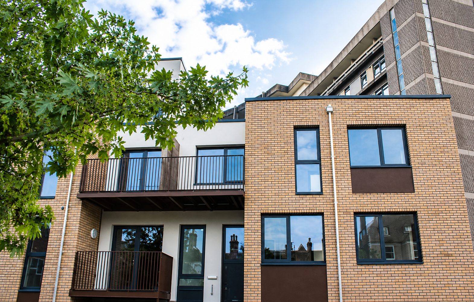 Buff brick slip beside a white-rendered balcony recess with black railings, a tree in the foreground