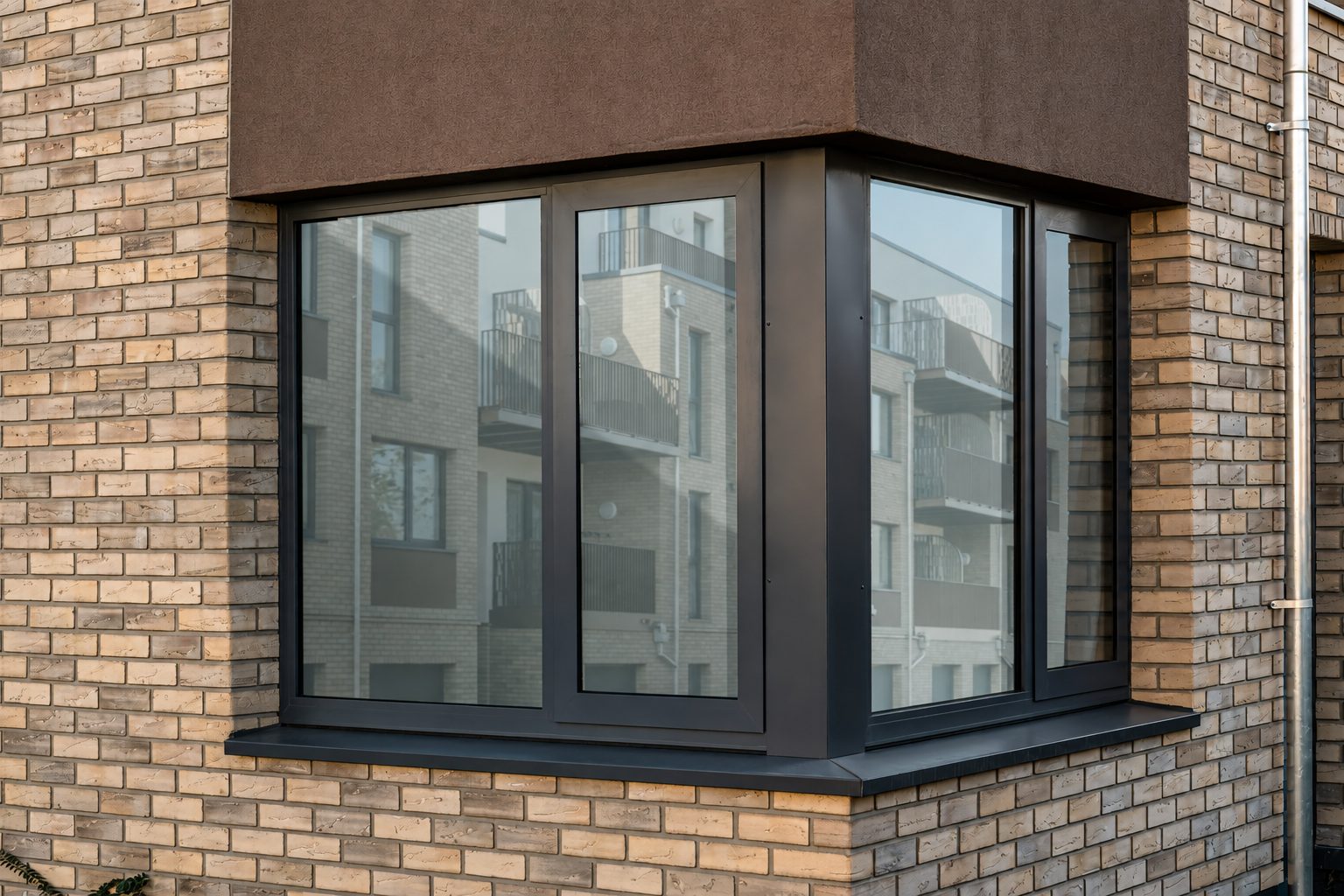 Close view of buff brick slips turning a corner around a dark-framed bay window, a brown rendered band above