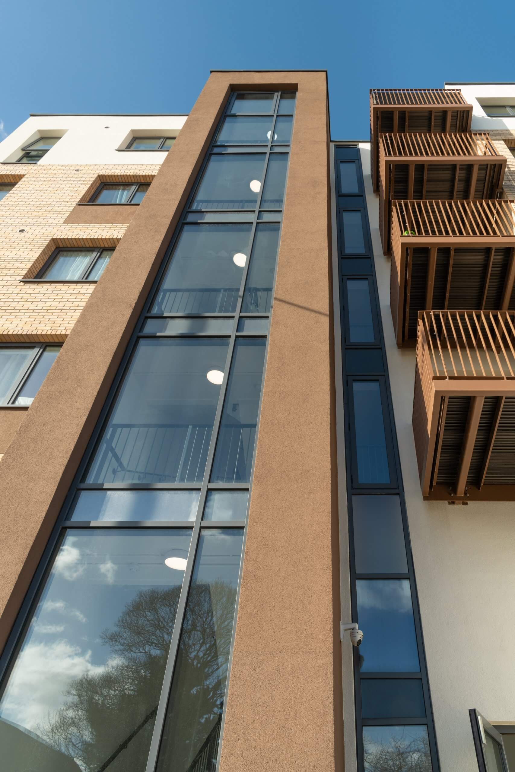 Looking up a tall glazed stair core in a brown rendered frame, brick slip to the left and timber-screened balconies to the right