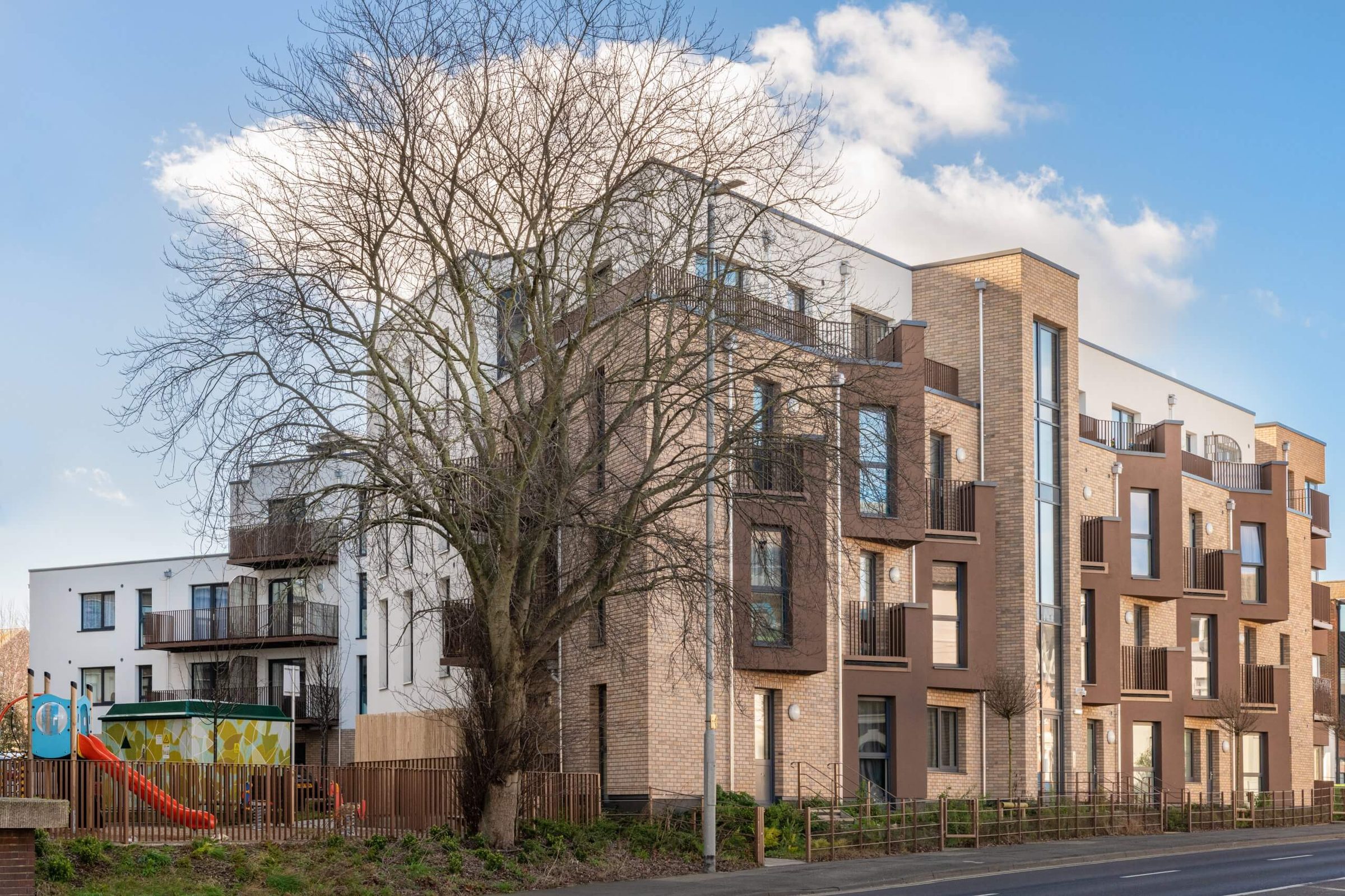 A buff brick-slip apartment wing with brown balcony surrounds and a glazed stair core along a road, a bare tree and a play area in front