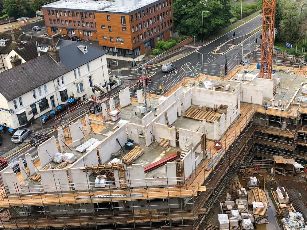 Aerial view of white block walls standing on a precast floor deck inside scaffolding, a red tower crane at the right