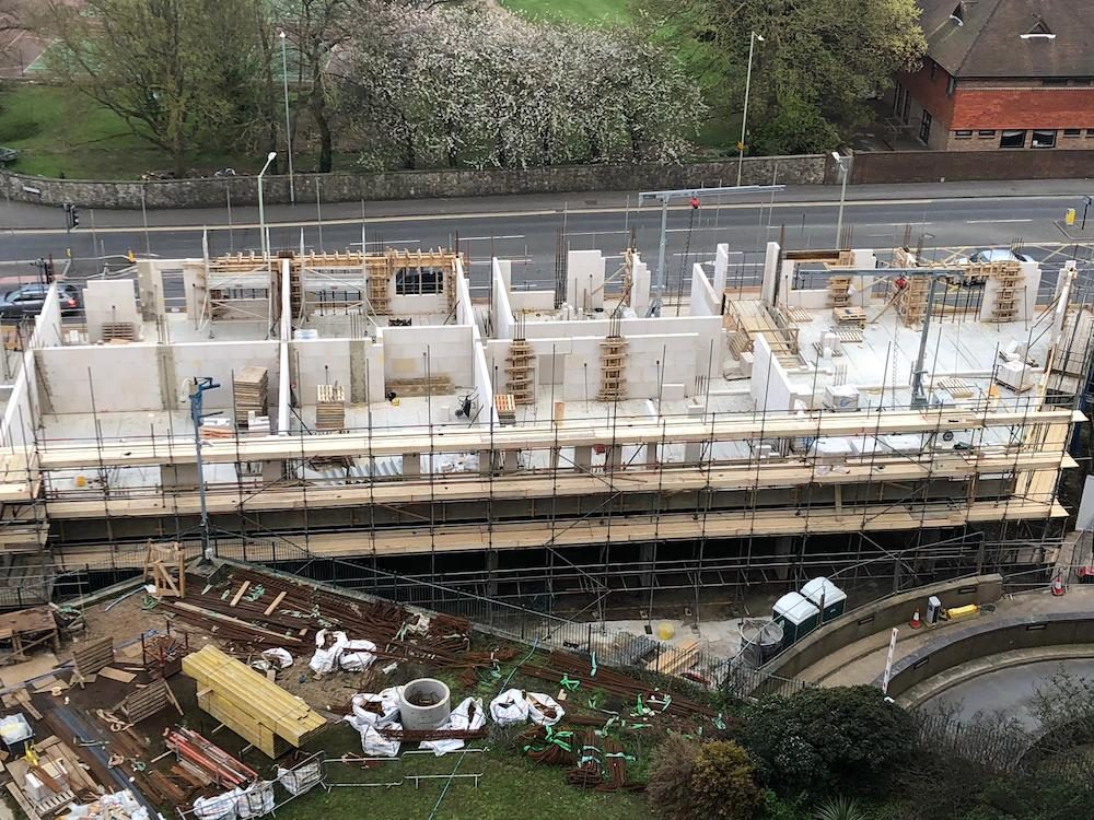 A floor of propped white block walls on a deck wrapped in scaffolding, a road and trees beyond