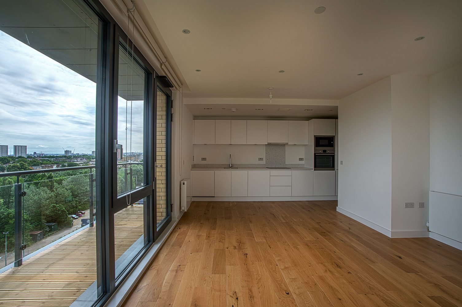 Empty apartment with an oak floor, a white kitchen and full-height glazing opening onto a balcony with trees and the city beyond