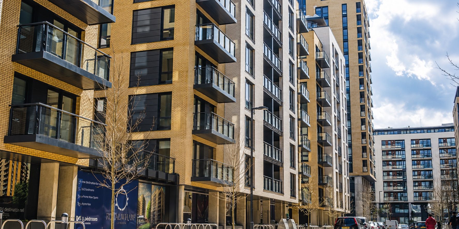 Street lined with buff and white brick residential blocks with dark projecting balconies, a blue sales hoarding at street level