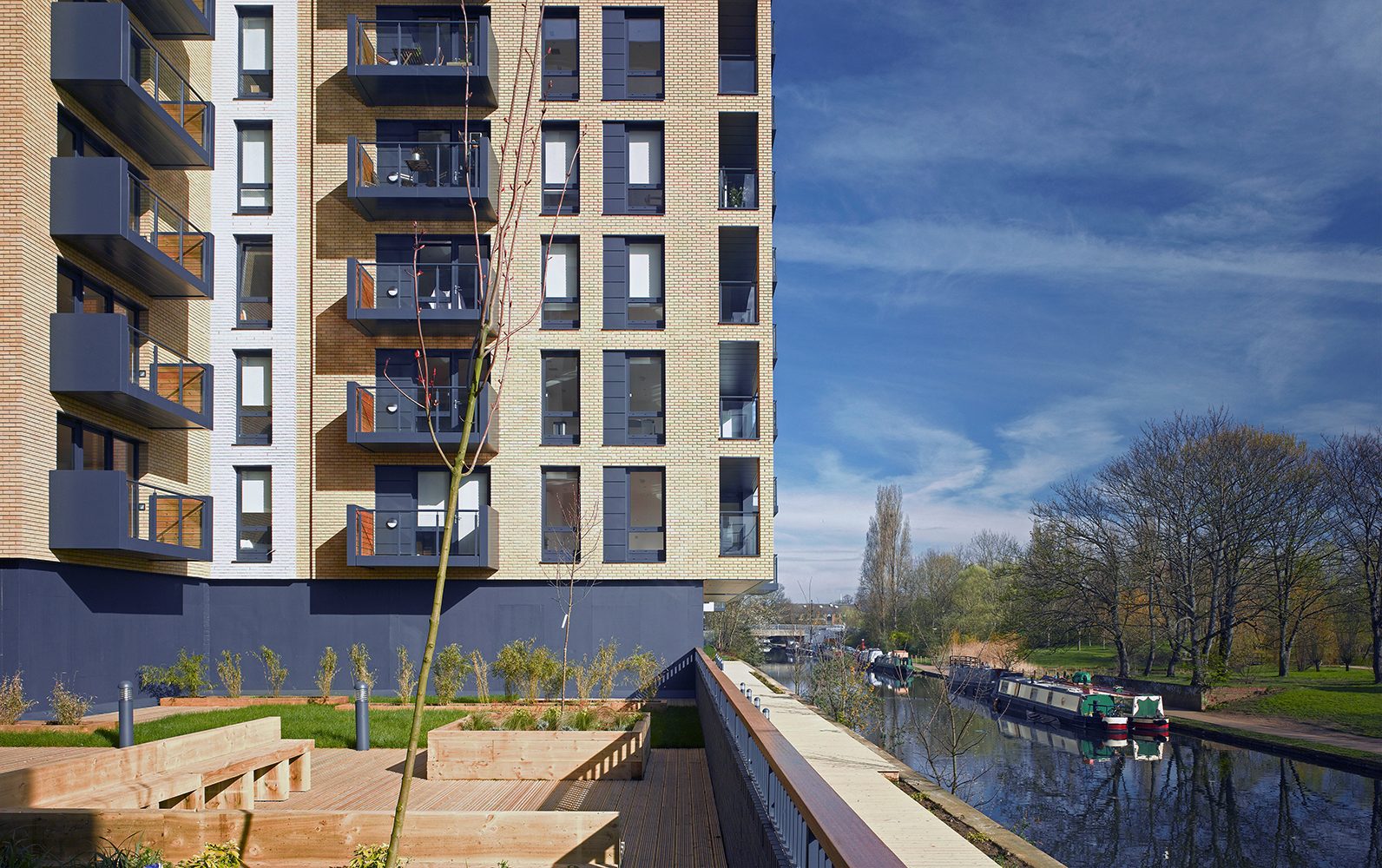Communal terrace with timber benches and planters beside a block with dark balconies, the canal and narrowboats running away to the right