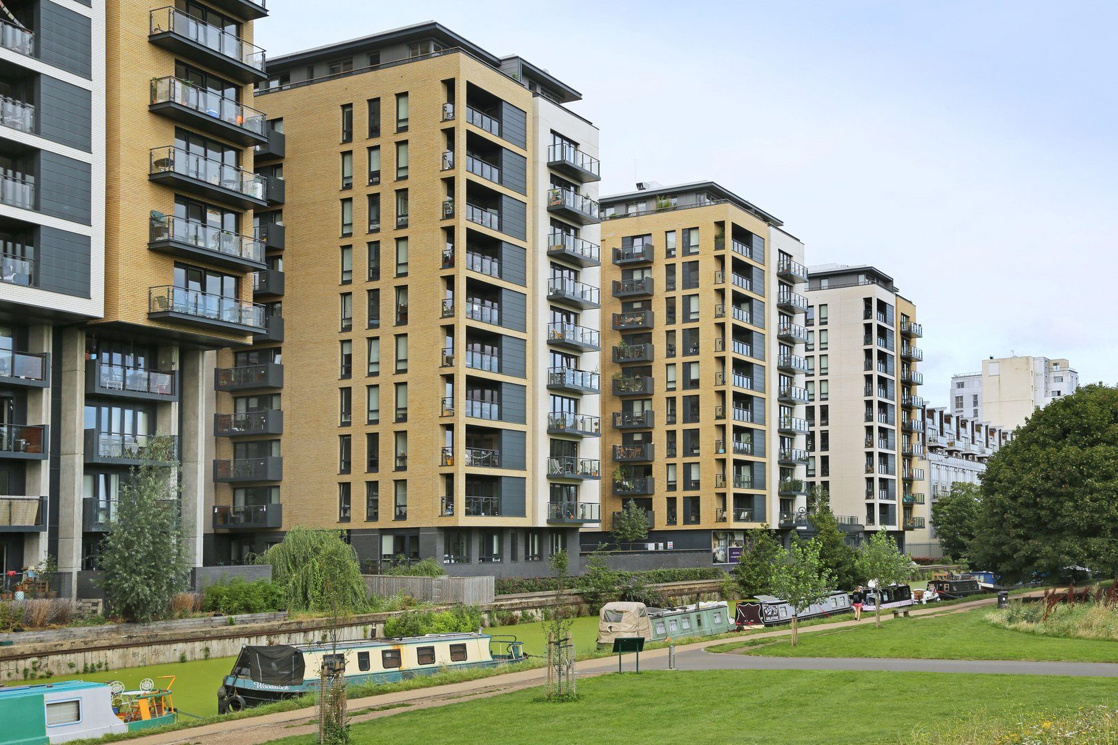Row of buff-brick residential blocks with grey and white panelled bays beside the Regent's Canal, narrowboats moored in front and a lawn in the foreground