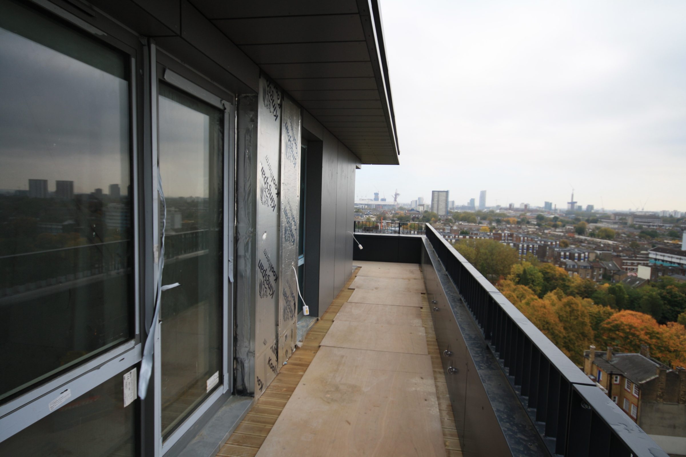 Penthouse terrace under construction with dark soffit and wall panels fixed, foil-faced Kingspan insulation board exposed beside a door, and east London beyond
