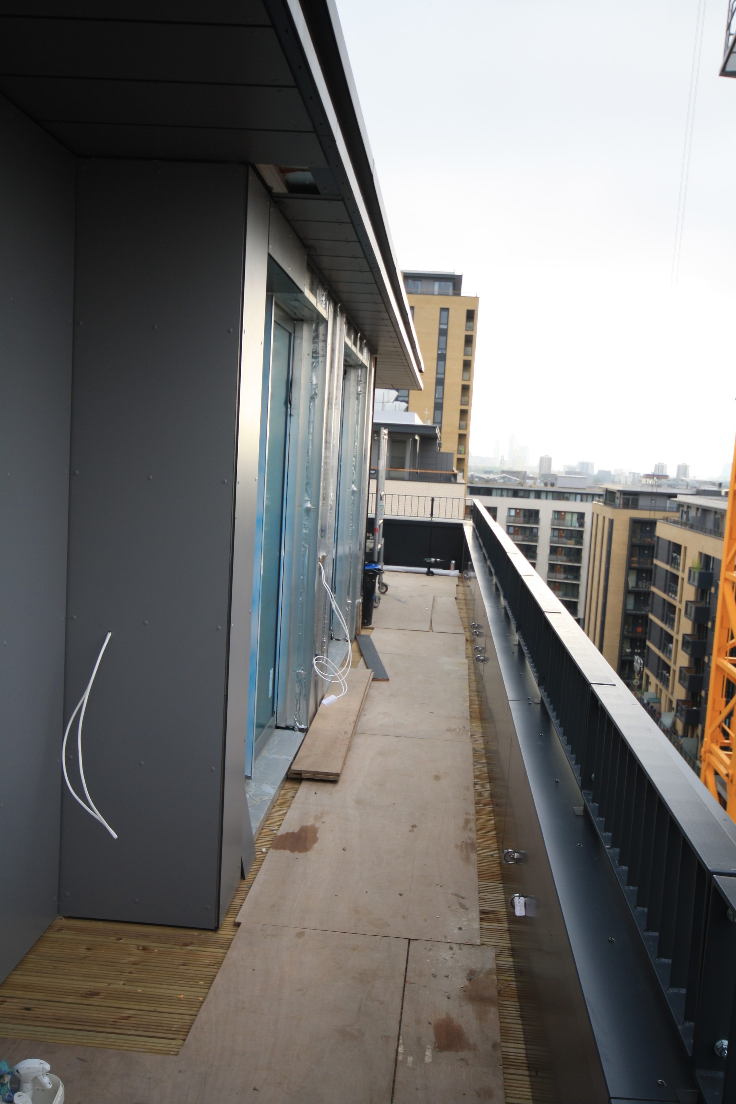 Penthouse terrace with dark full-height wall panels, a panelled soffit above and neighbouring residential blocks and a tower crane beyond