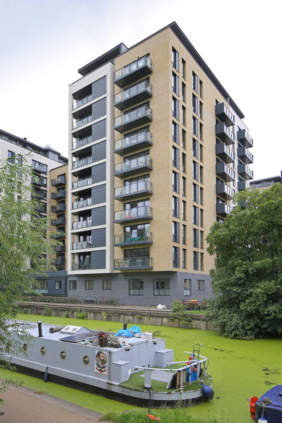 Residential tower with grey panelled bays and buff brick piers rising above a duckweed-covered canal with a moored barge