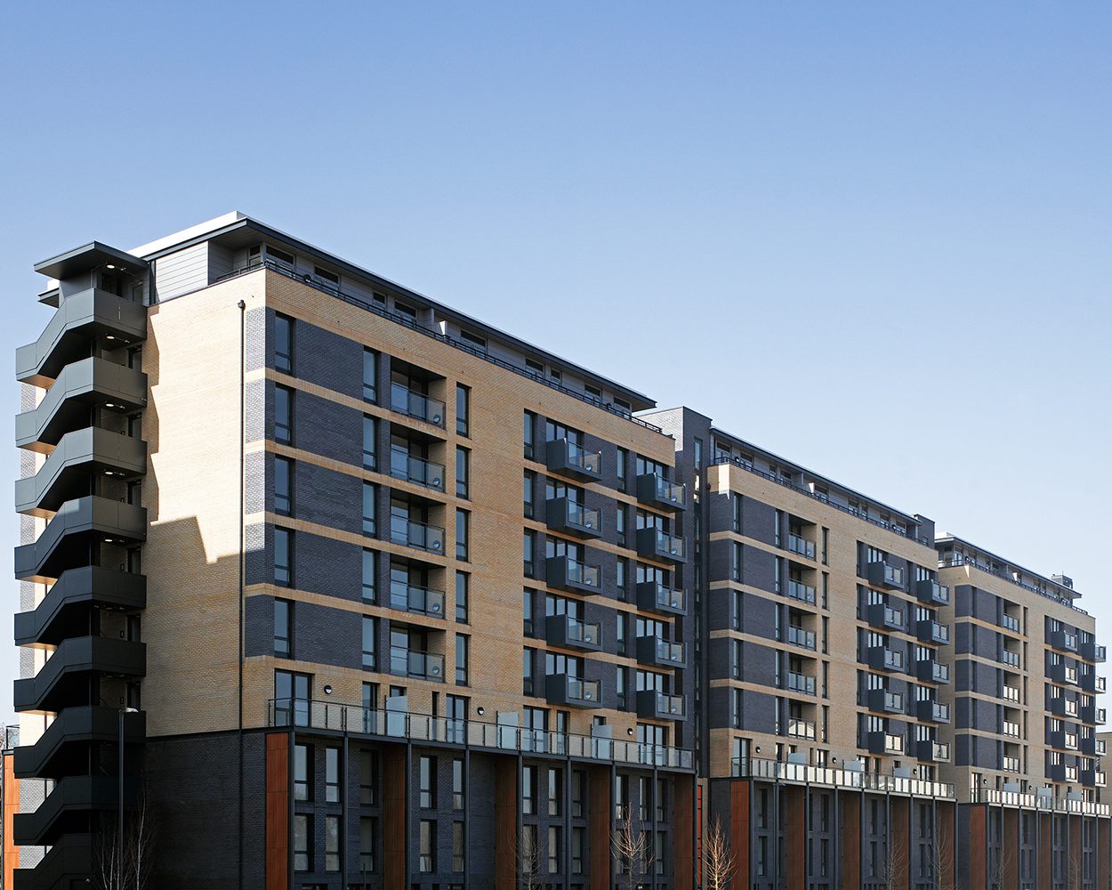 Long residential elevation in buff brick with dark panelled bays, a dark brick base and a stack of projecting balconies at the corner