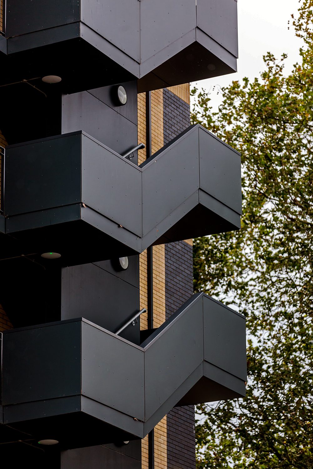 Dark panelled projecting balconies stacked one above another, each folding around a corner, against buff brick and trees