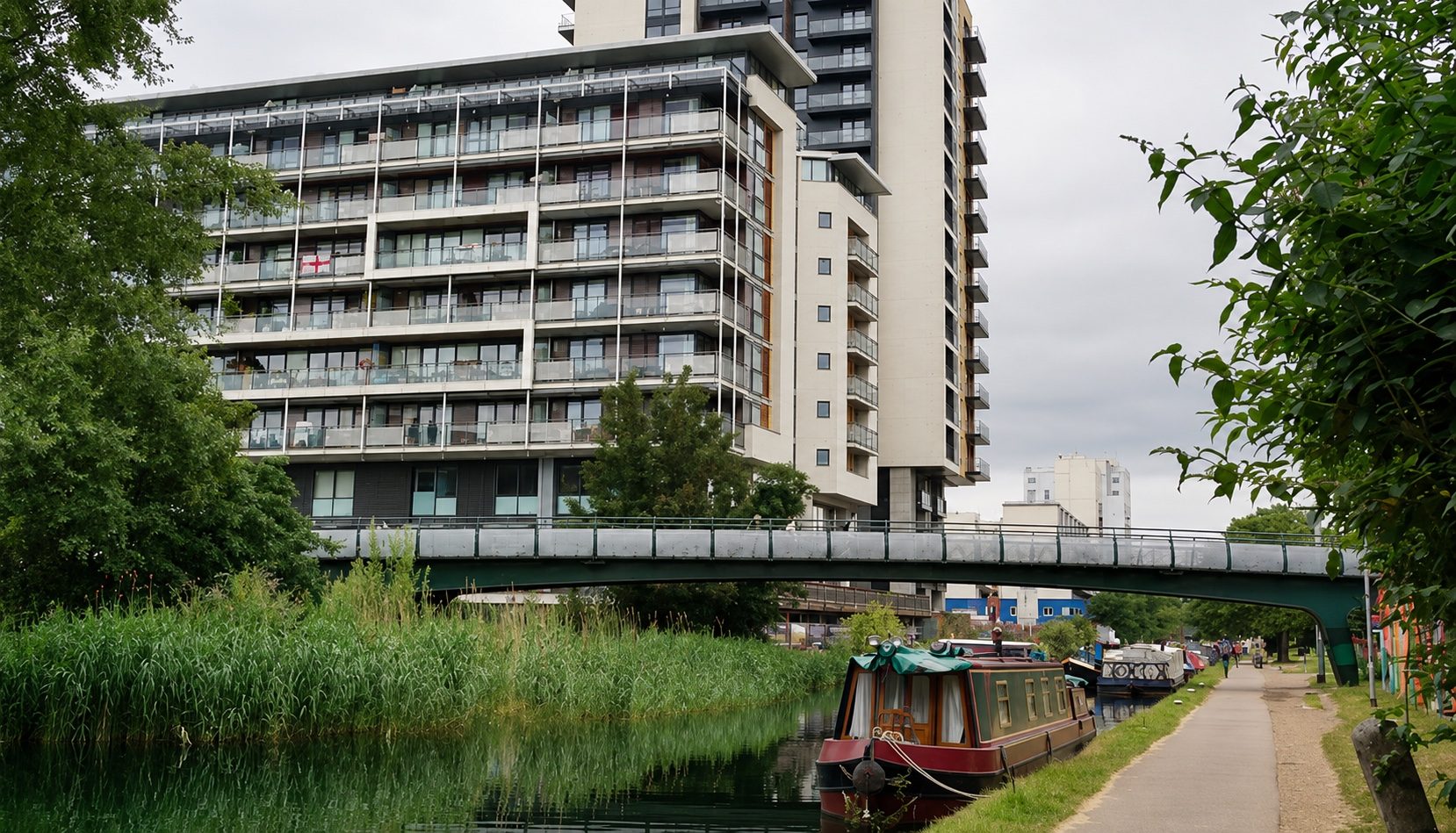 Canal towpath with a moored narrowboat and a footbridge, residential blocks rising behind the trees