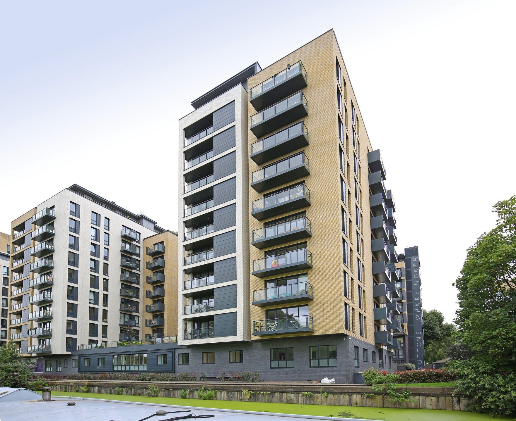 Buff-brick tower with a grey panelled bay and glass balconies above a planted canal bank, a vertical Suttons Wharf North sign on the block to the right