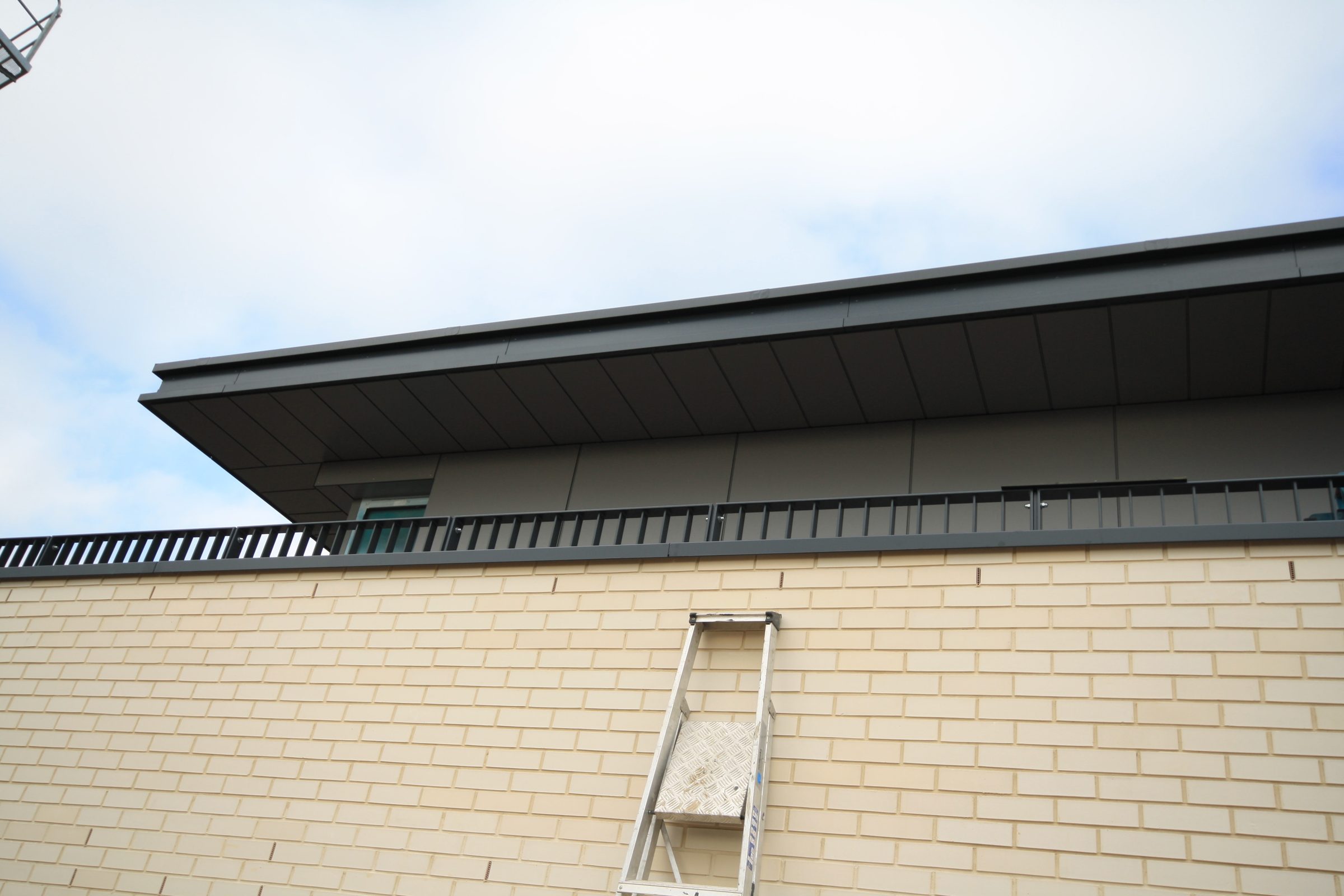 Dark top-floor overhang and soffit above a railing and a buff-brick parapet wall, a ladder leaning against the brick