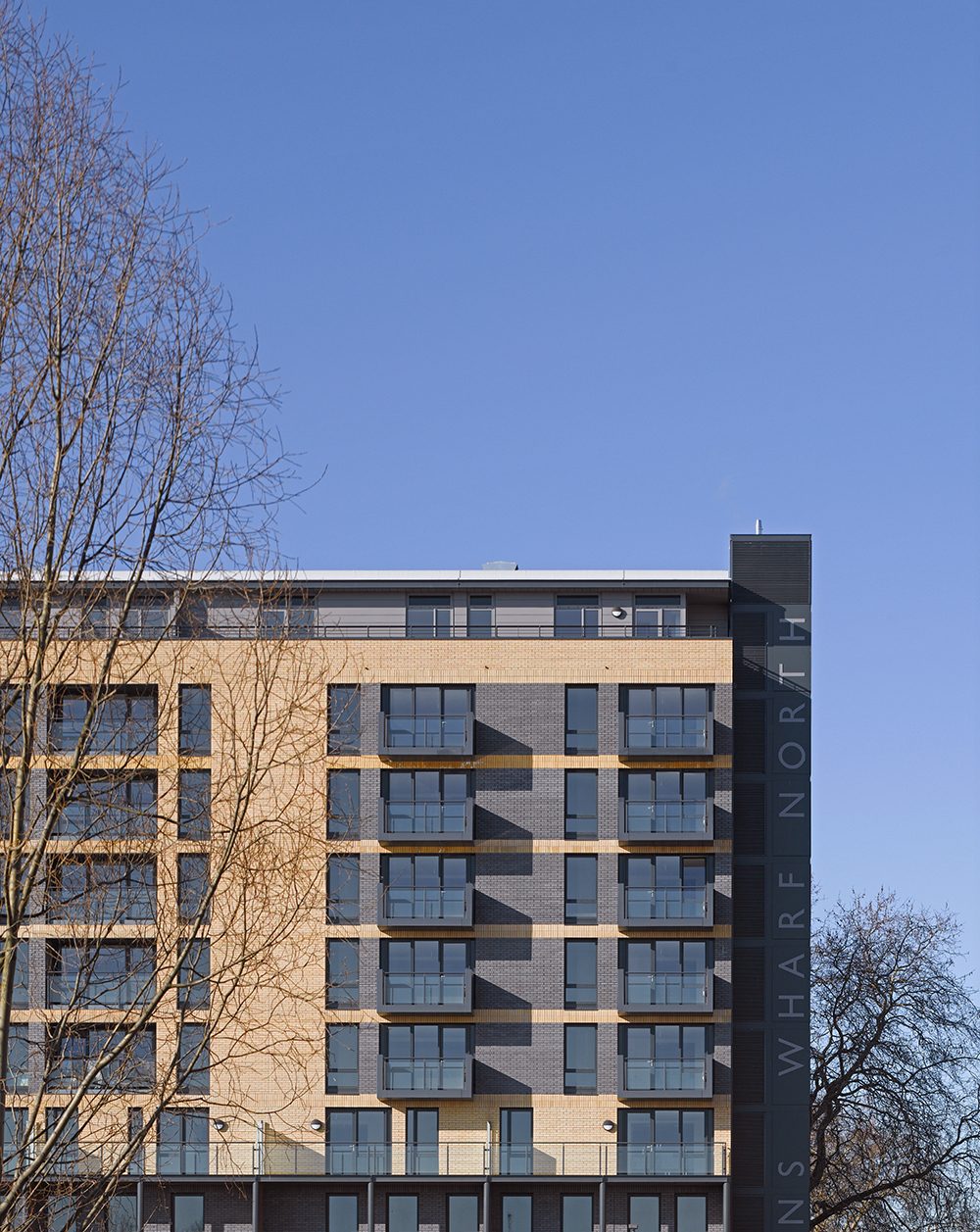 Buff-brick block with grey panelled bays and balconies, a tall dark sign reading Wharf North at its corner, bare trees either side